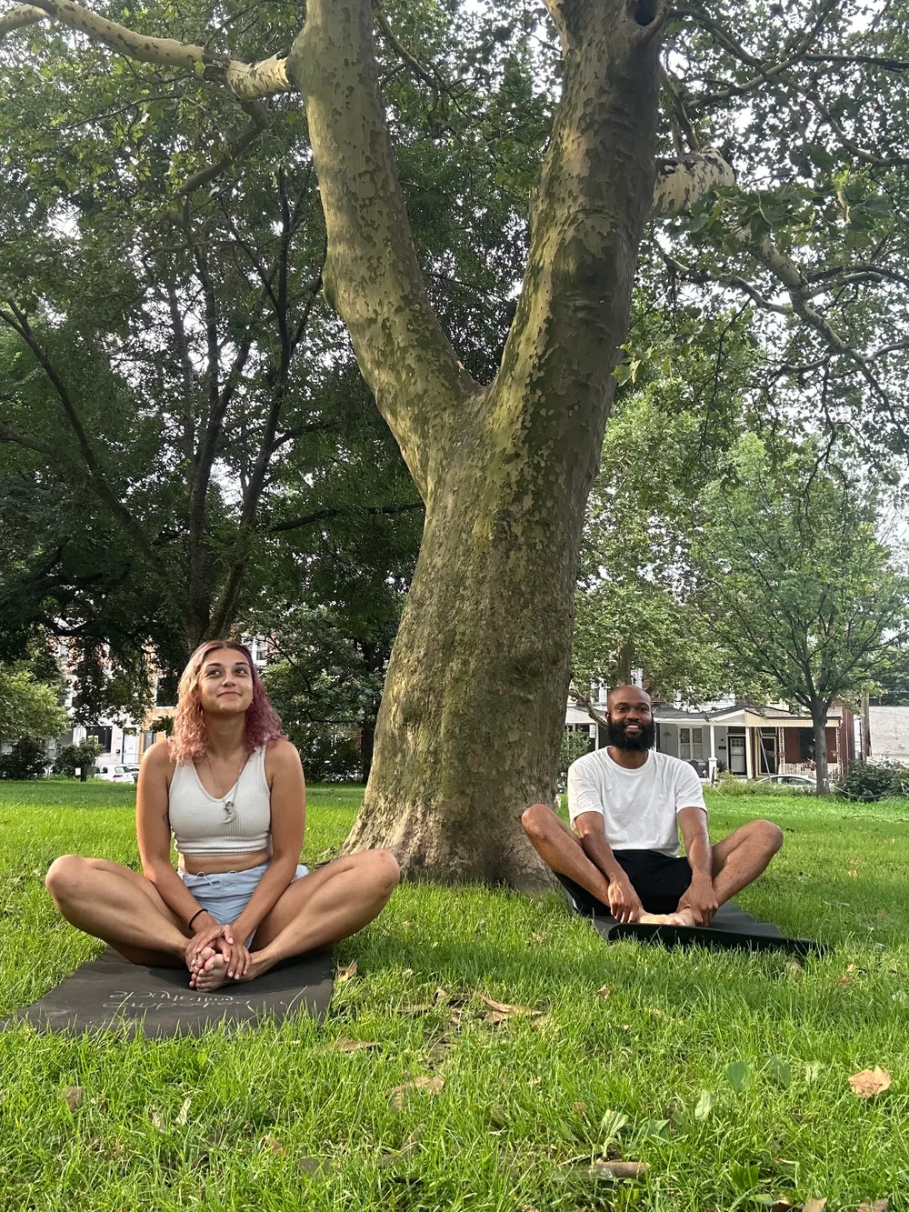 Two people sit cross-legged in the grass by a tree during the day, looking peaceful