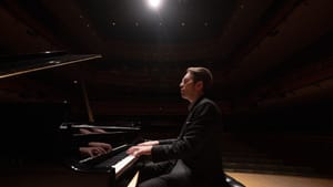 Andsnes, a white man with brown hair wearing a black suit, is seen in profile playing a grand piano on the Perelman stage.