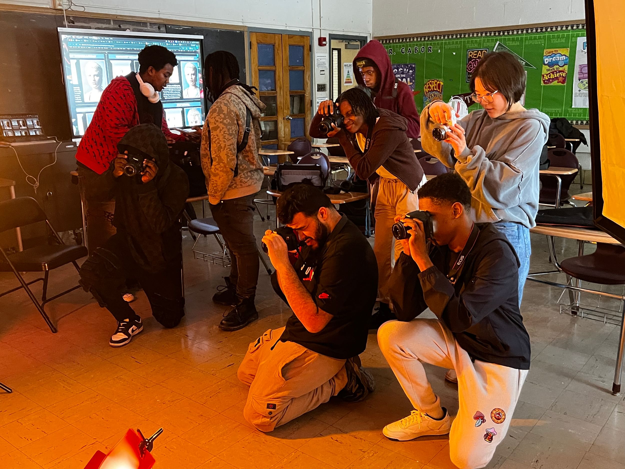 A group of teenagers and young adults each have a handheld camera, aiming at something off image, in a classroom setting