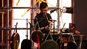 Chang playing a wind instrument with percussion instruments around him on stage, a silhouetted crowd before him