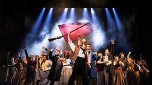 About two dozen actors in 19th-century costumes pose under dramatic spotlights and fog, each raising one fist in the air.