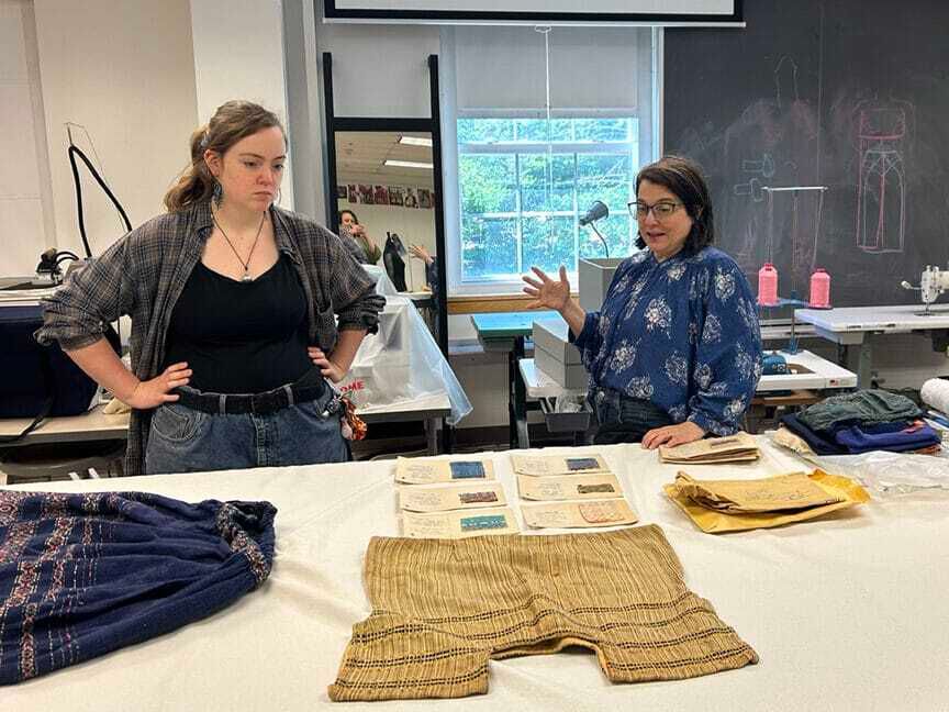 Two white women in a bright daytime studio talk to each other while looking at Esherick textiles laid on a white table.