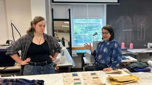 Two white women in a bright daytime studio talk to each other while looking at Esherick textiles laid on a white table.