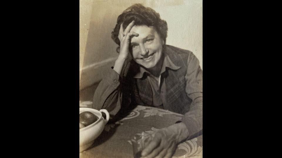 Black & white photo of Esherick, a white woman with short dark curly hair, smiling & leaning on a table, a drink in one hand
