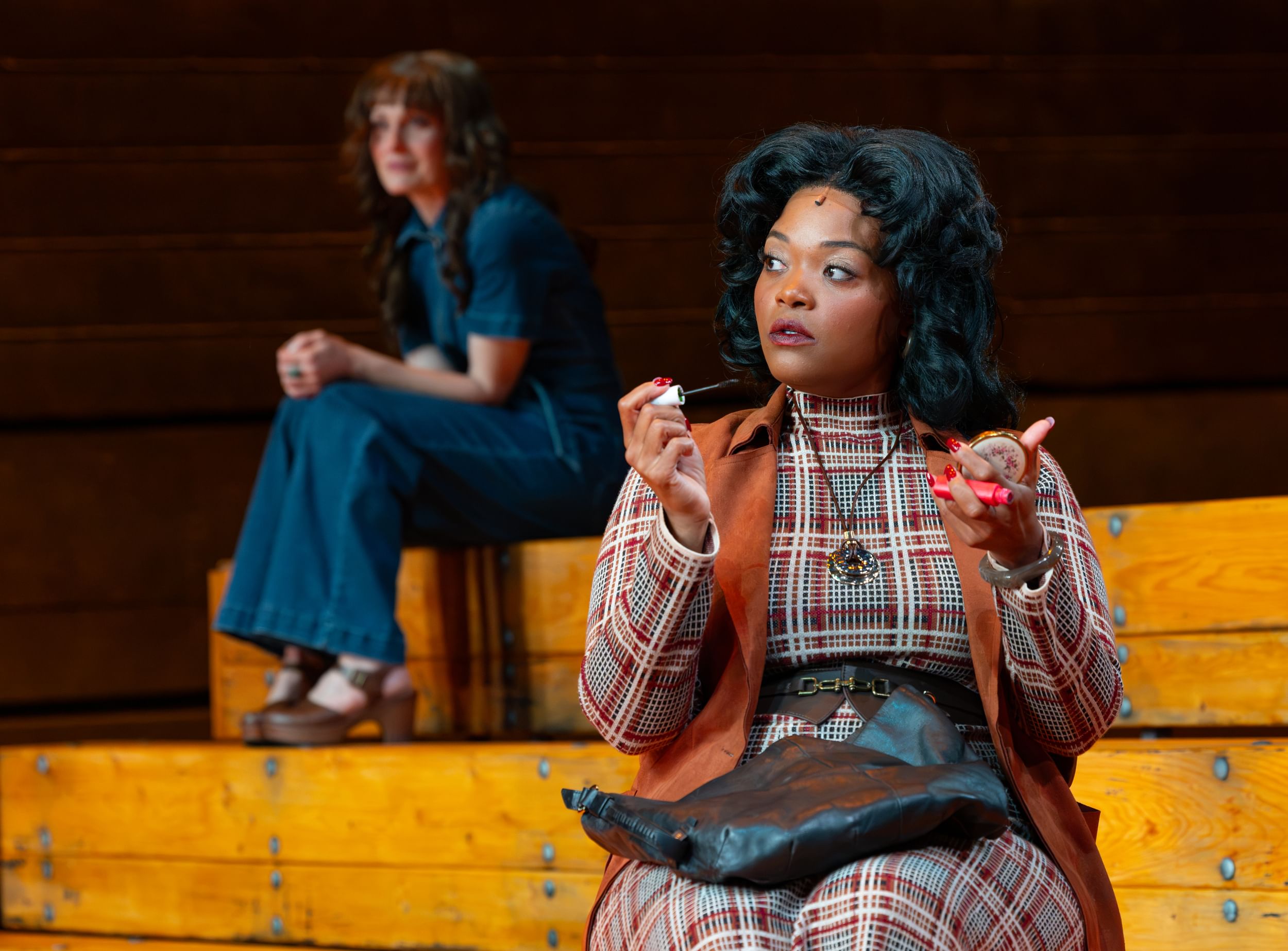 Johnson, sitting and applying makeup in a brown plaid dress, looks warily to the side while Zinkel sits in the background.