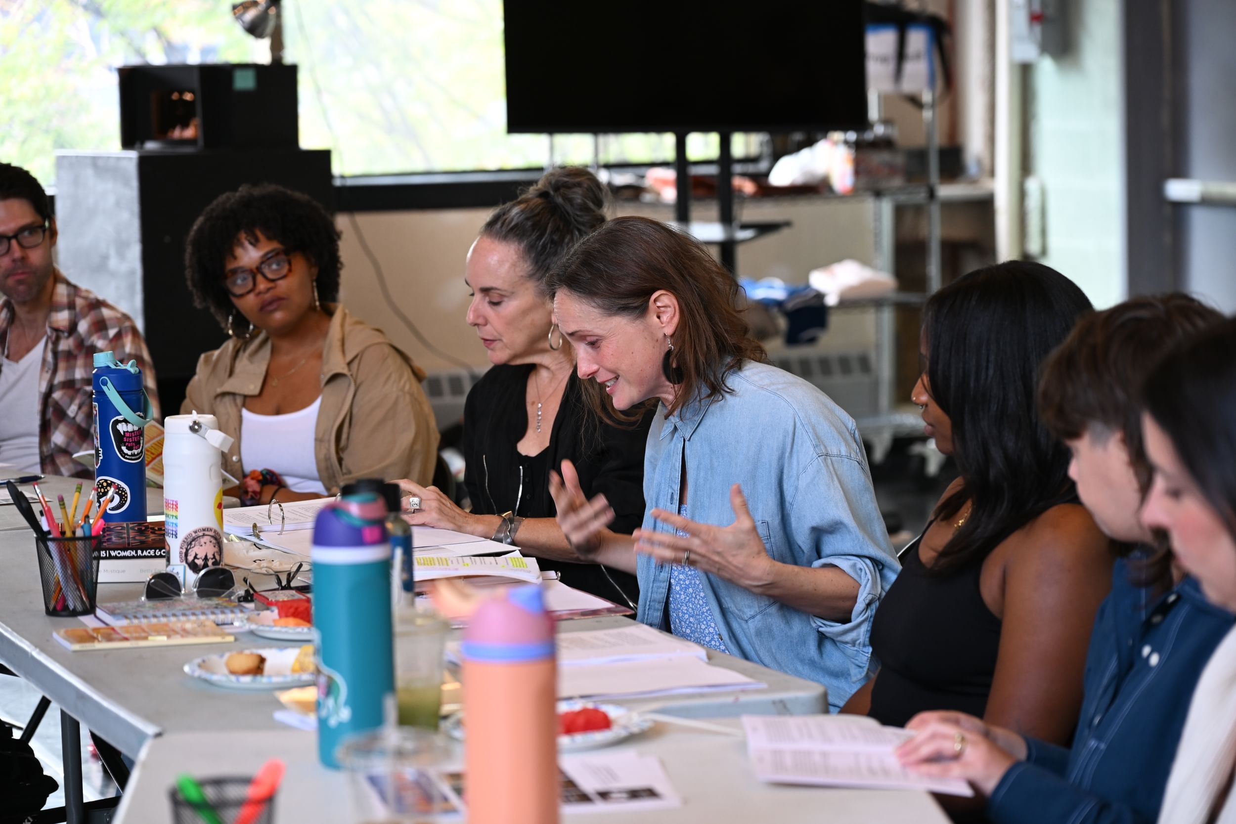 Seven people in casual clothes around a table crowded with scripts, water bottles, and pens focus on a reading.