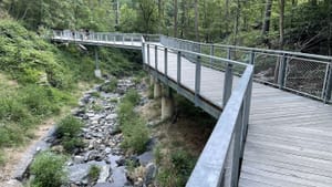 A bridge with flat gray boards and metal railings in angled sections crosses a stony brook. Three people walk on it.