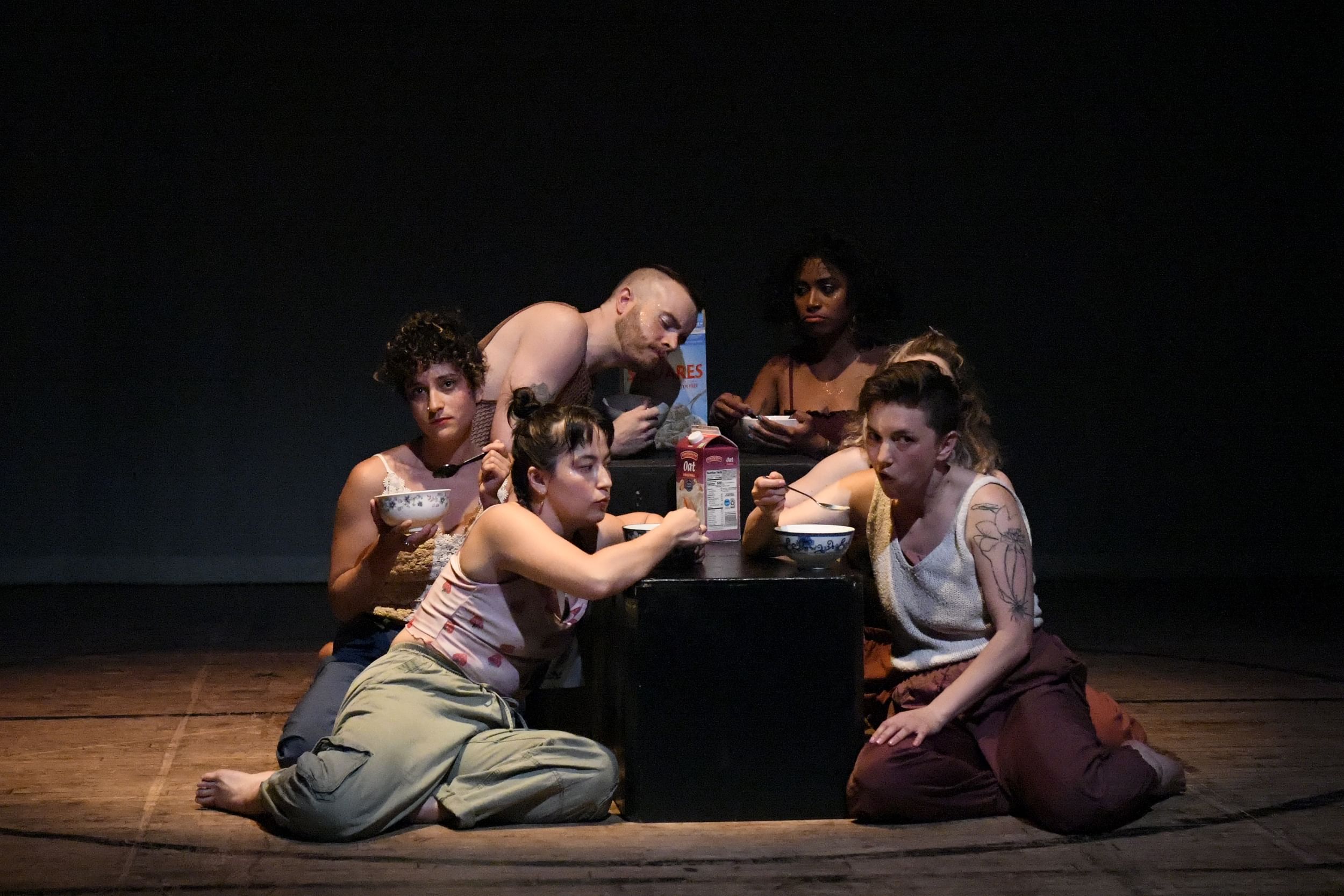 Six dancers of different races and genders sit slouch on the floor around a table of boxes, pretending to eat morning cereal