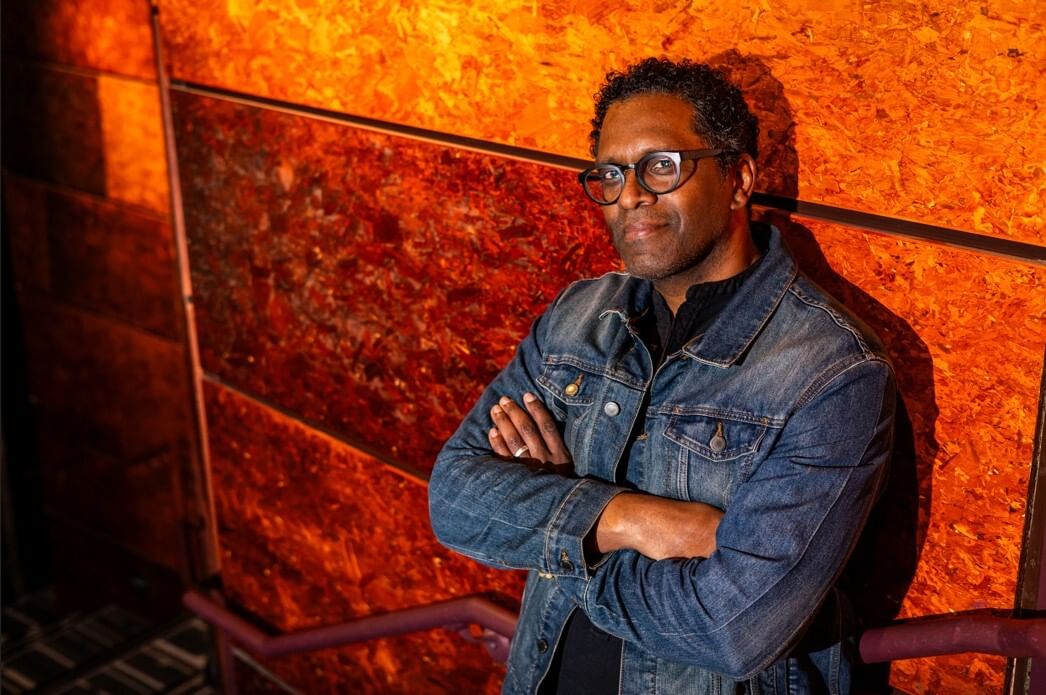 Lindsay, a Black man, leans against a textured orange wall with his arms crossed, wearing glasses and a denim jacket.