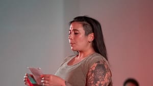 Lubar, a white woman wearing a gray tank top, her arm tattooed with an eagle and flowers, reads from a piece of paper.