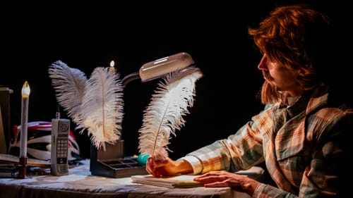 Close-up on Yorke in a plaid coat, stick-on beard and brown wig, writing with a big white ostrich-style quill pen.