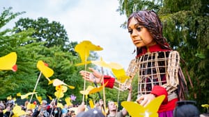 A 12-foot-tall puppet girl with a wooden frame reveals someone piloting inside. A crowd surrounds Amal in a park area