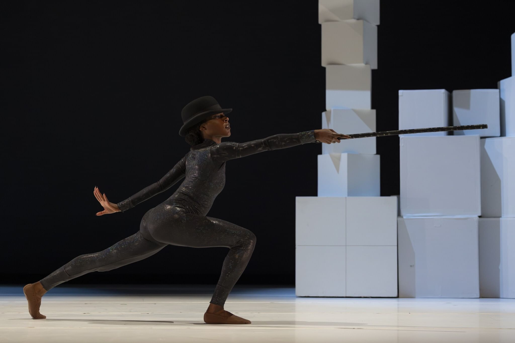 Simpson, a Black woman in an iridescent black bodysuit and bowler hat, poses in profile with a knee bent, arms outstretched