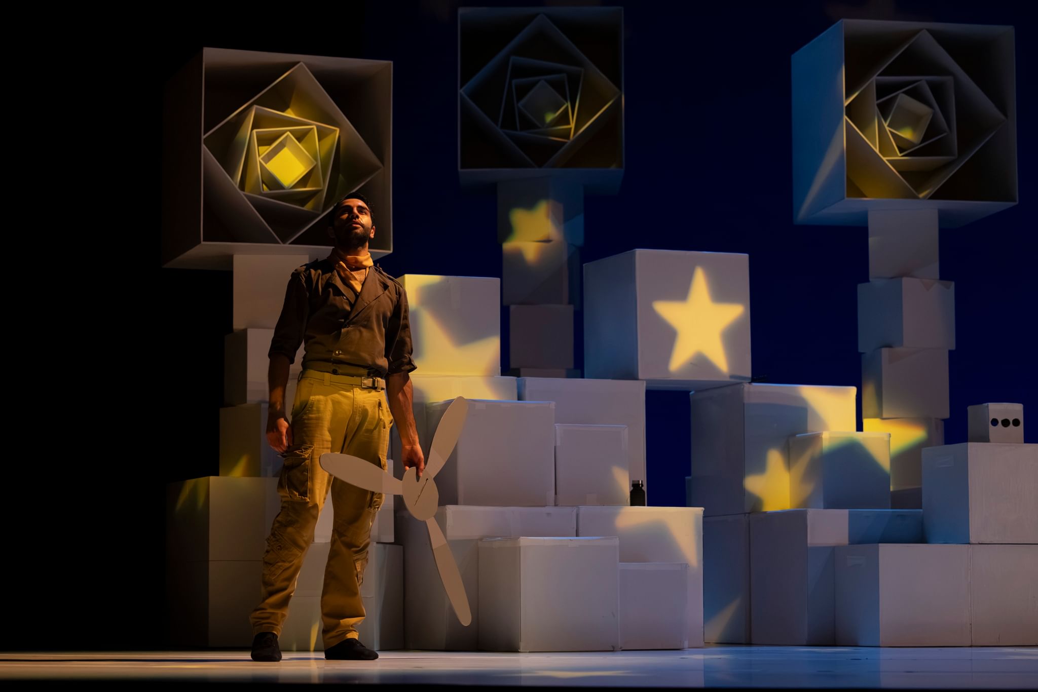 Montepara, in fatigues and brown shirt, stands holding a propellor in front of stacked white boxes with yellow stars of light