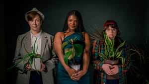 Three people stand next to each other facing camera holding house plants against a dark backdrop