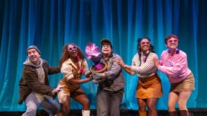 In 60s or 90s fashion, the cast mugs happily in a line, reaching out to each other. Lieberman holds a purple plant at center.