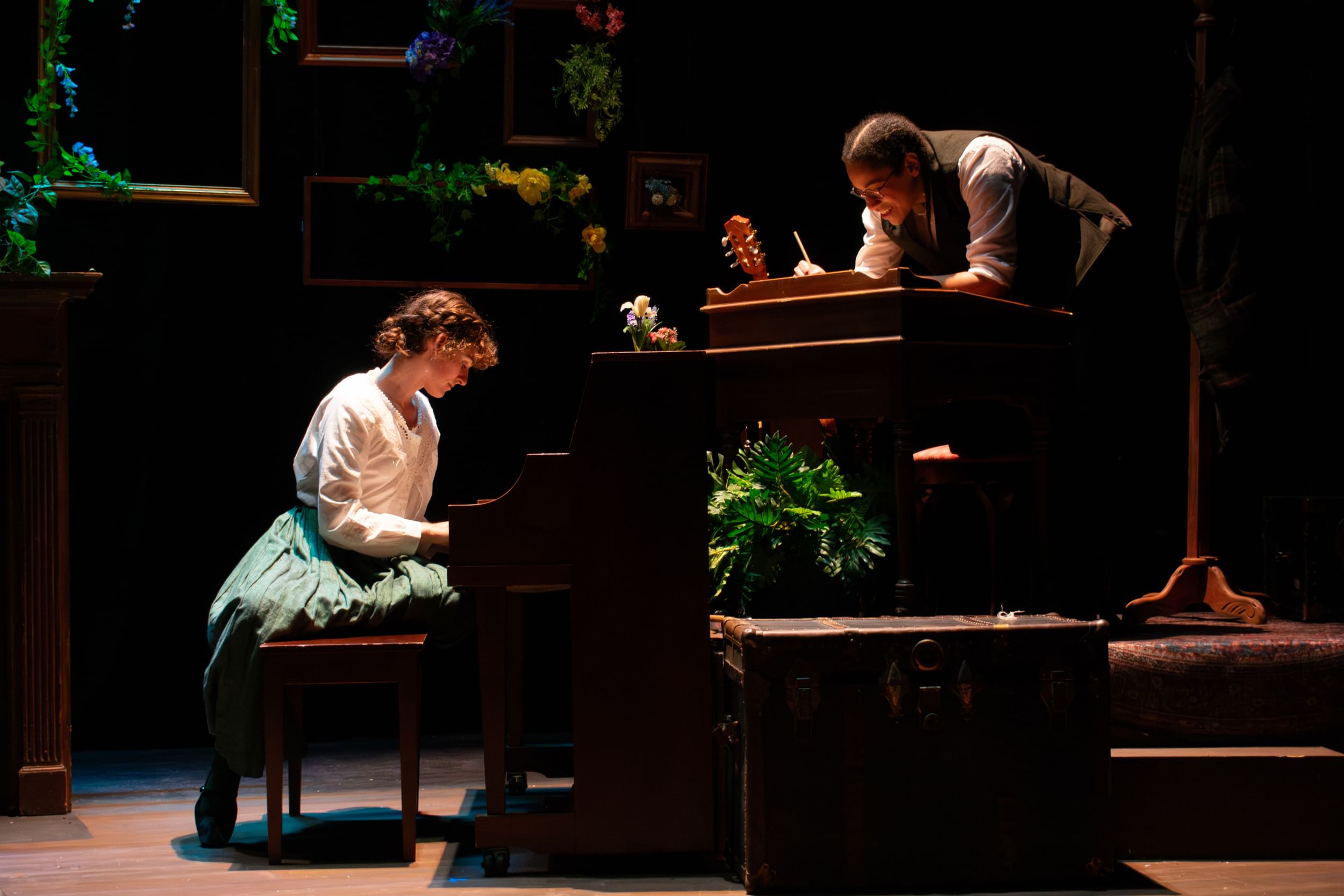 Two women around a piano on stage. One sits at the piano playing the keys, another is at a desk close by writing