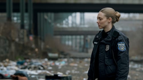 Seyfried, a white woman with blond hair wearing a police uniform, stands in an outdoor area full of trash.