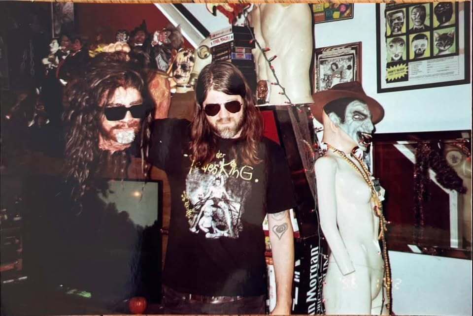 Longo holds up a prop head that looks like his, wearing glasses and long hair, a variety of horror film props around him