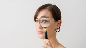 Portrait shot of Loots, in a cream-colored dress, holds a magnifying glass up to her left eye
