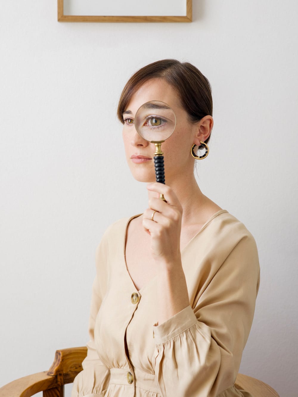 Portrait shot of Loots, in a cream-colored dress, holds a magnifying glass up to her left eye