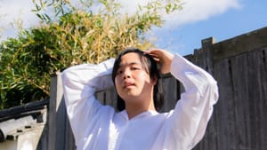 Liyou poses with hands near their head, in a voluminous white outfit. They're outdoors below a blue sky with white clouds.