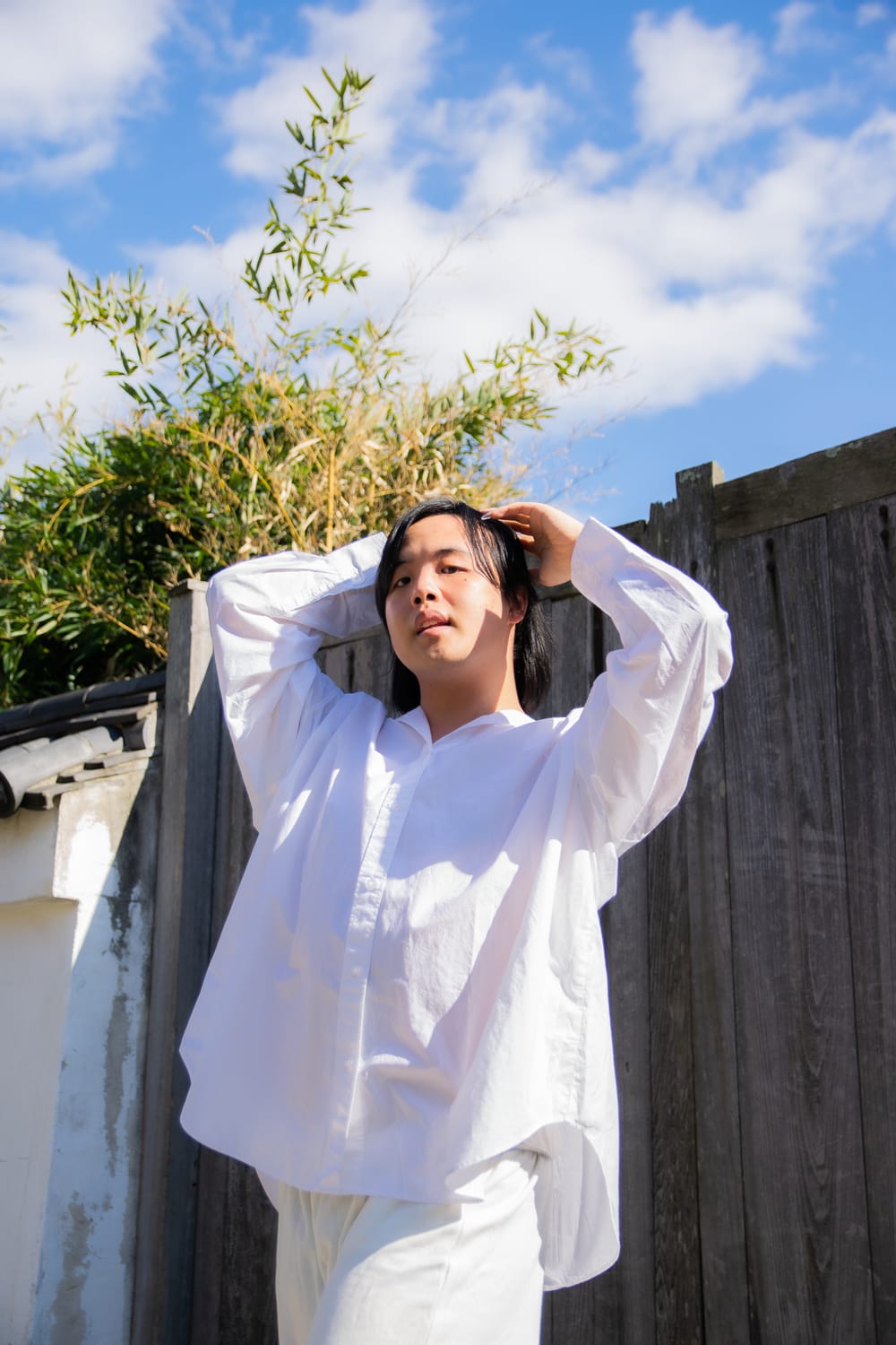 Liyou poses with hands near their head, in a voluminous white outfit. They're outdoors below a blue sky with white clouds.