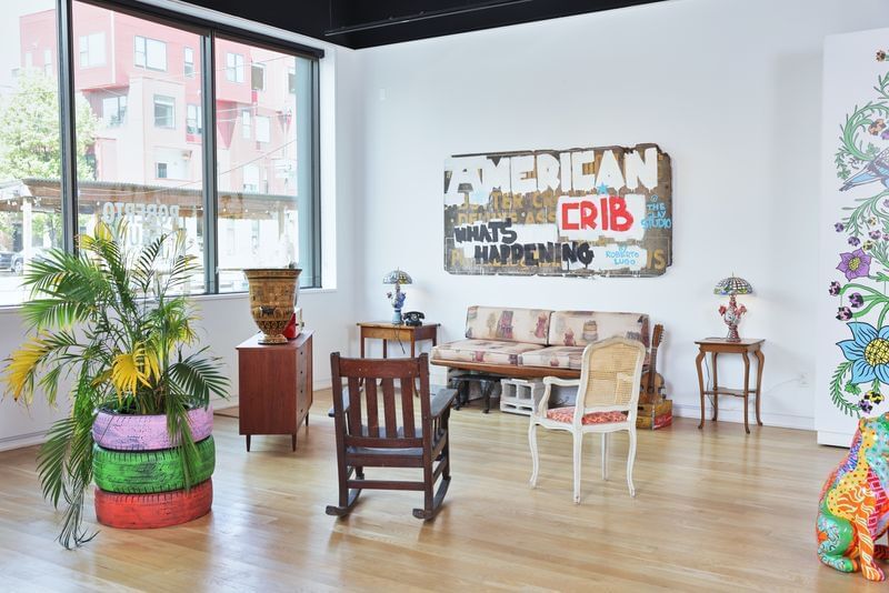 Sunny white gallery with wooden floors & large windows has eclectic furniture, Lugo lamps, and fern growing in stacked tires