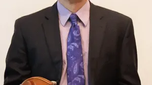 Mazzocchi sits in a black suit with a blue tie, holding a violin for a portrait style photo.