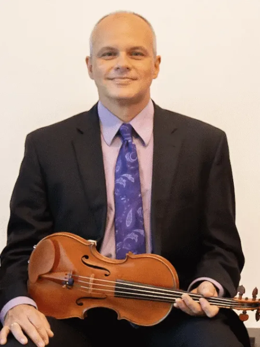 Mazzocchi sits in a black suit with a blue tie, holding a violin for a portrait style photo.