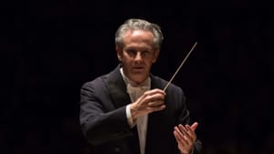 Close-up on the conductor, with gray hair, glasses, and a black tux conducting with a baton, with darkness behind him.