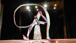 A woman of Asian descent in a white silk dress with blue and floral accents weaves long cloth streams across a large stage