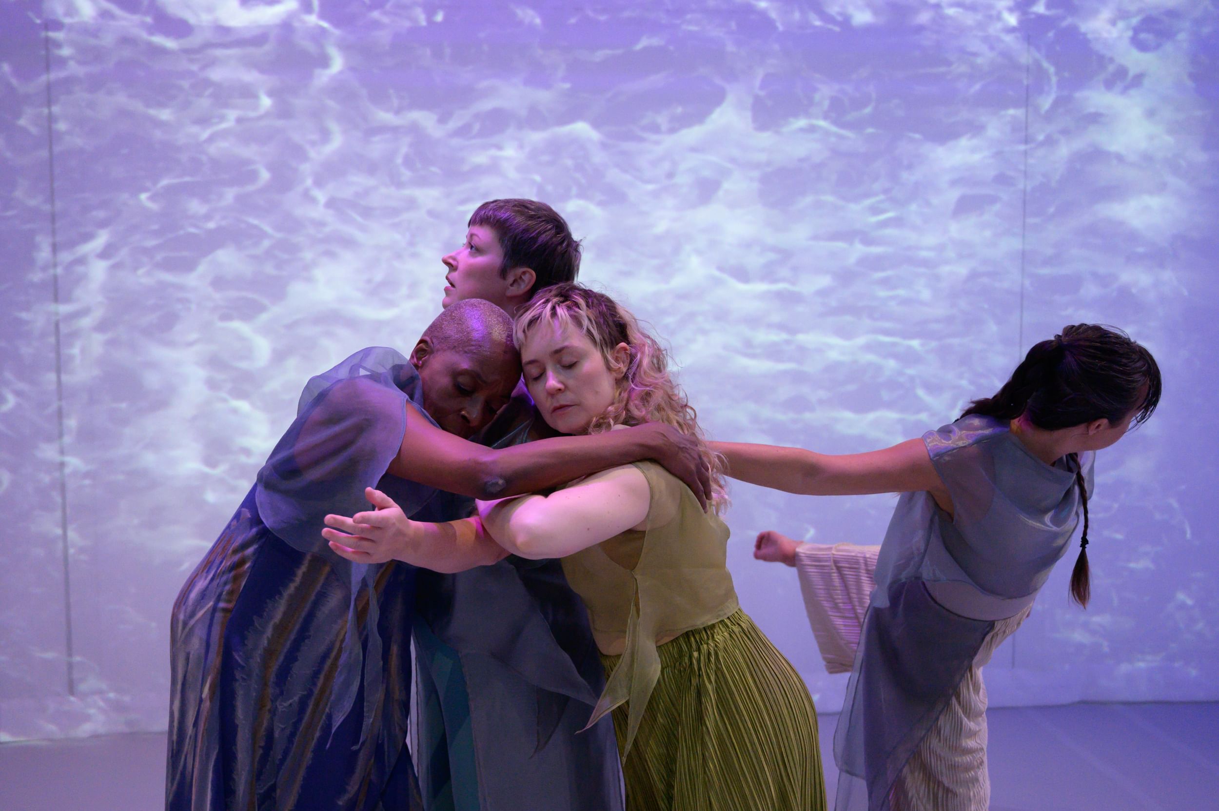 Four diverse dancers interact in a dreamy, sea-like setting, three embracing tenderly and one leaning away.