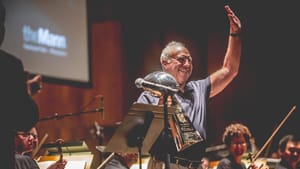 Lurie grins, holding the giant metallic trophy and waving with the other hand, orchestra musicians around him onstage.