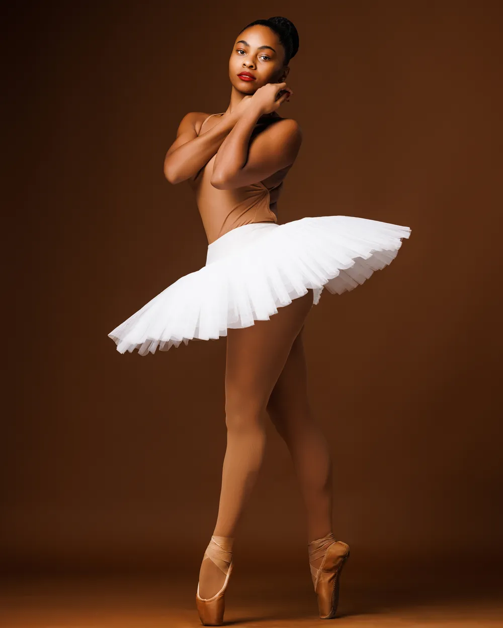 A young Black woman dancer in a skin-matched leotard & tights and white tutu poses en pointe.