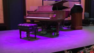 The Lyric Fest stage at Longwood Gardens, with a piano under purple lights. Fern-like plant with red flowers line the stage.