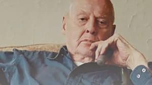 Rorem, an elderly white man in a blue button-down, reclines contemplatively in a chair, cane in one hand.