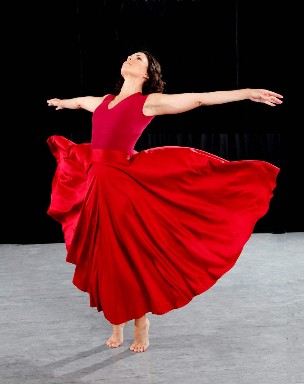 A dancer in a red dress that flows out wide, her arms stretched out long, on a stage with a black background