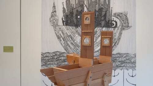 A large wooden sculpture that evokes a boat or a creature made with fine wood planks and a pen & ink illustration behind