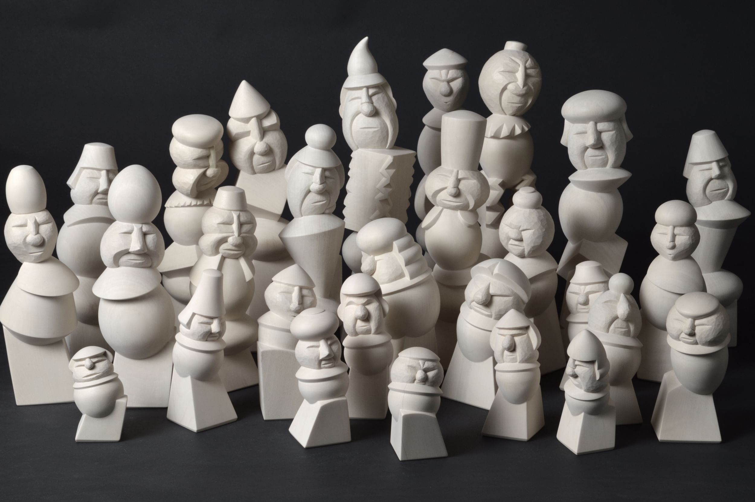 About 30 small white wooden figurines of fantastical yet familiar people on tiny pedestals, on a black background