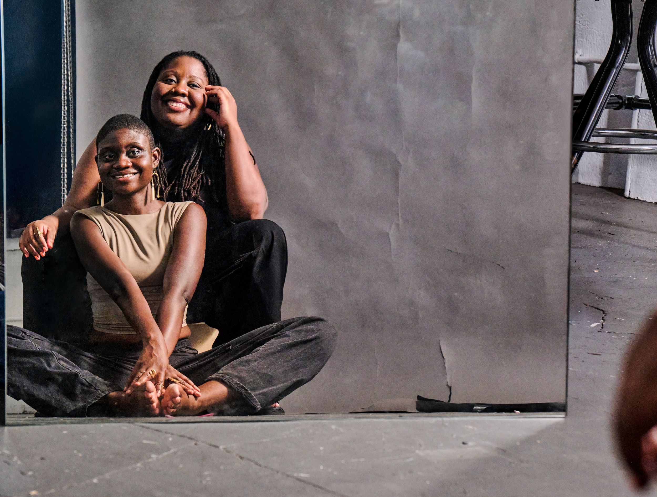 Two Haitian American women sit facing a mirror, one person in front of the other, smiling joyfully, in portrait style photo