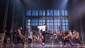 A large dance ensemble surrounding him, Markus strikes a signature pose, one leg extended with white shirt fluttering behind