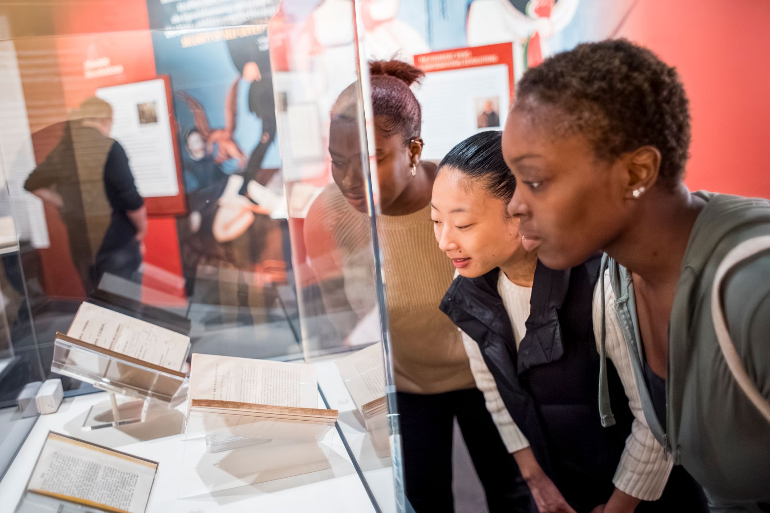 Two Black people and one Asian person look closely at historic books enclosed in a museum vitrine.