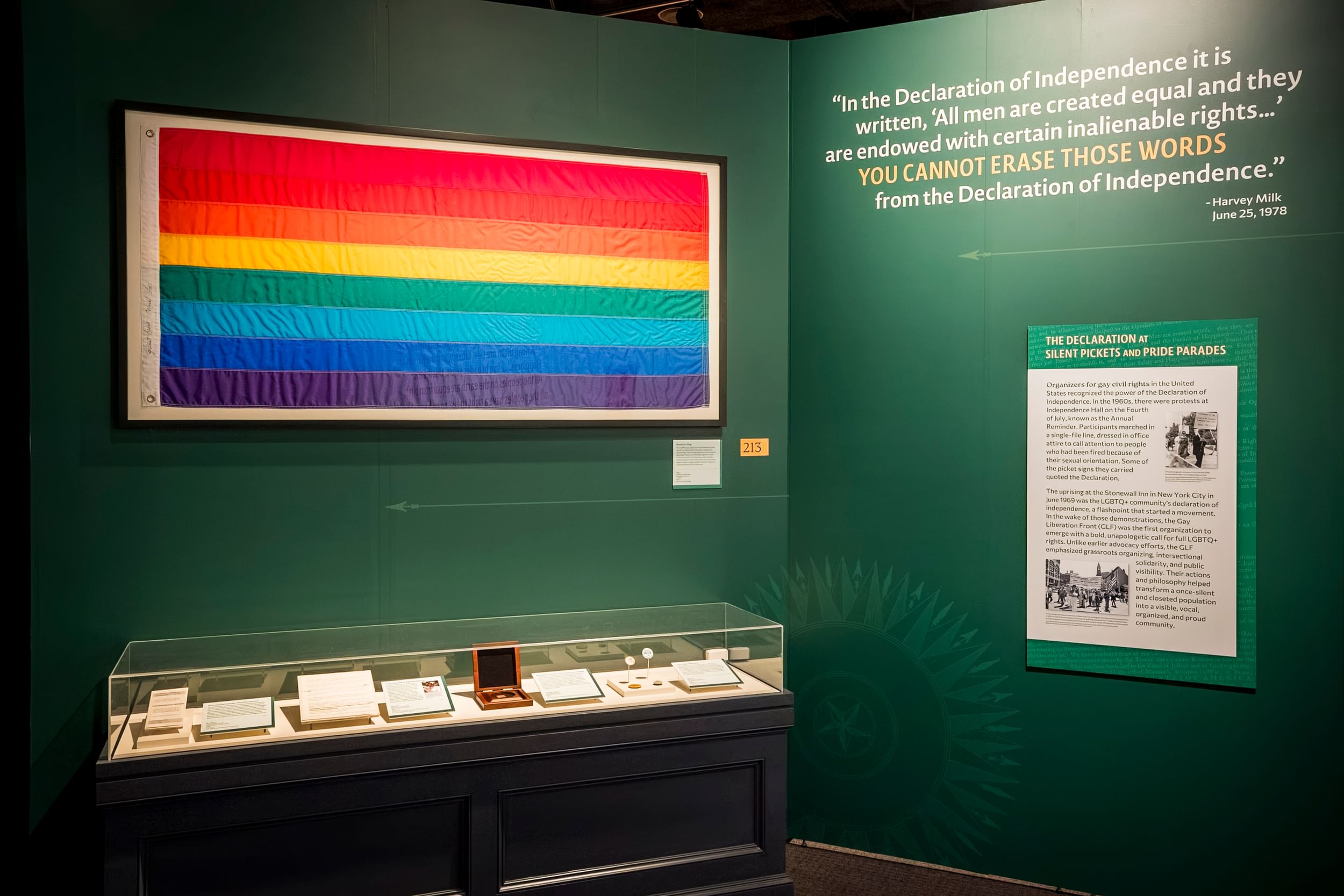 In a frame on a green gallery wall, an original version of the rainbow Pride flag over a vitrine showing historic objects.