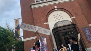 The museum’s boxy brick façade, with white trim. In front, a protestor holds a sign reading “this museum hosts facists”