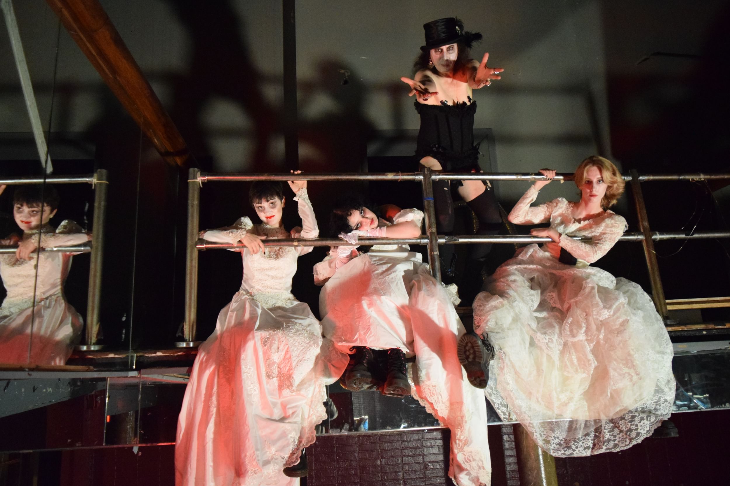 Three women in ragged pink princess dresses hang from a mezzanine, below Rudy in a corset and top hat, reaching forward