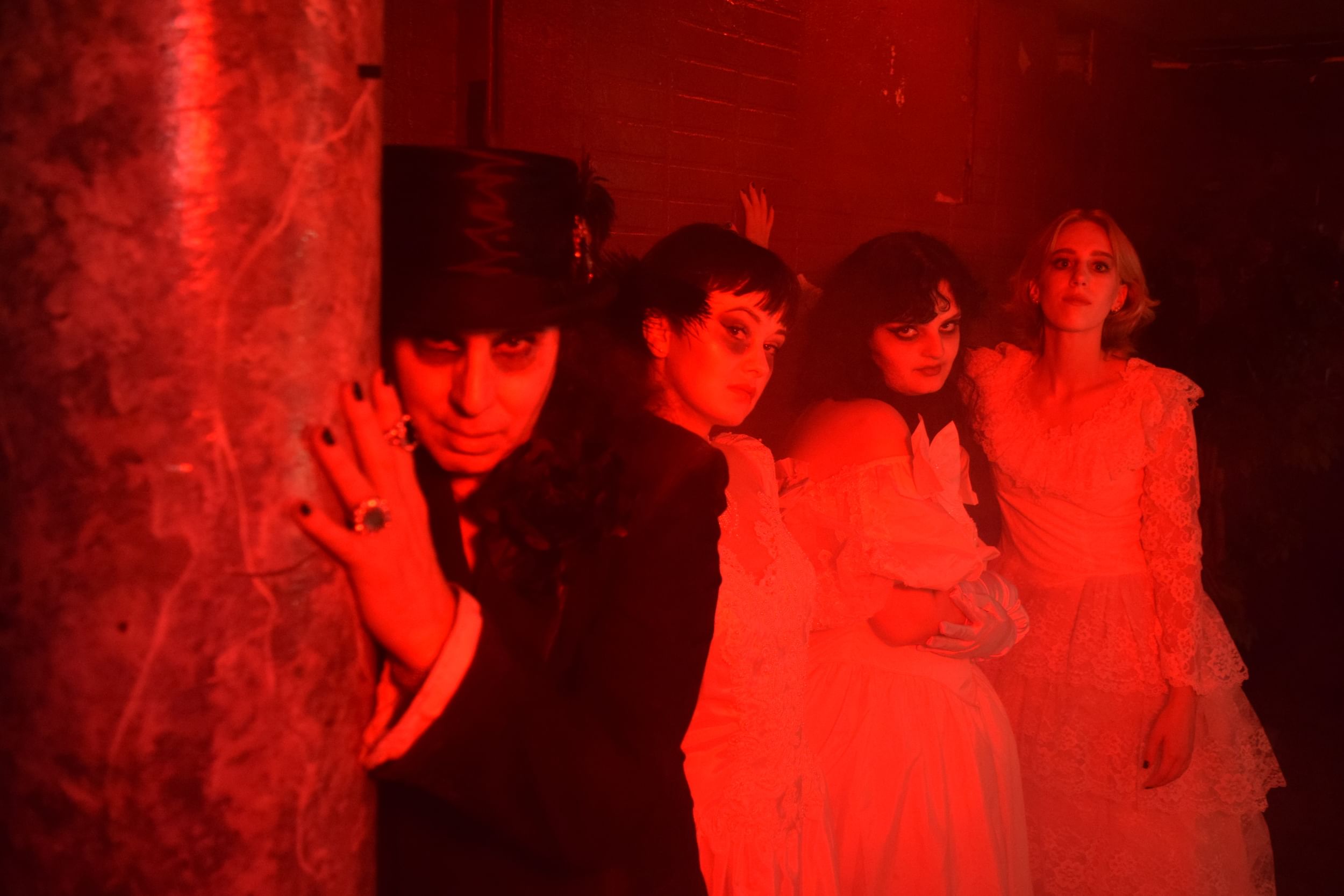 In dramatic red light, Caporaso lurks behind a pillar with the 3 brides behind him. They all wear smudged black eye makeup.