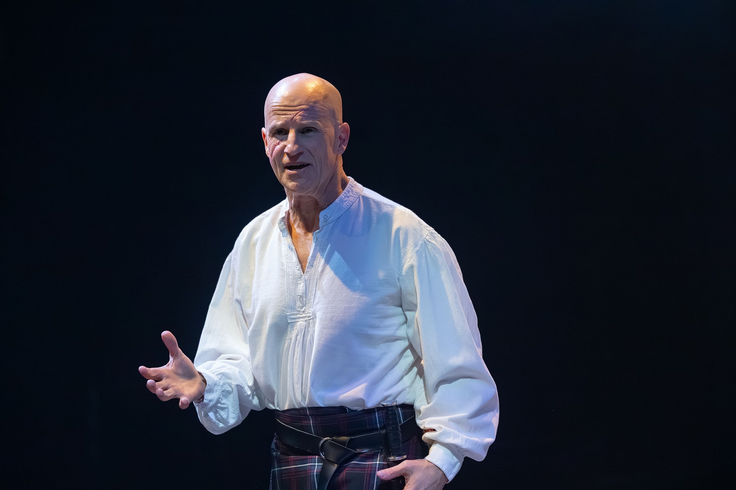 Lawton, a bald white man in a loose white shirt and striped kilt, gestures while he speaks.