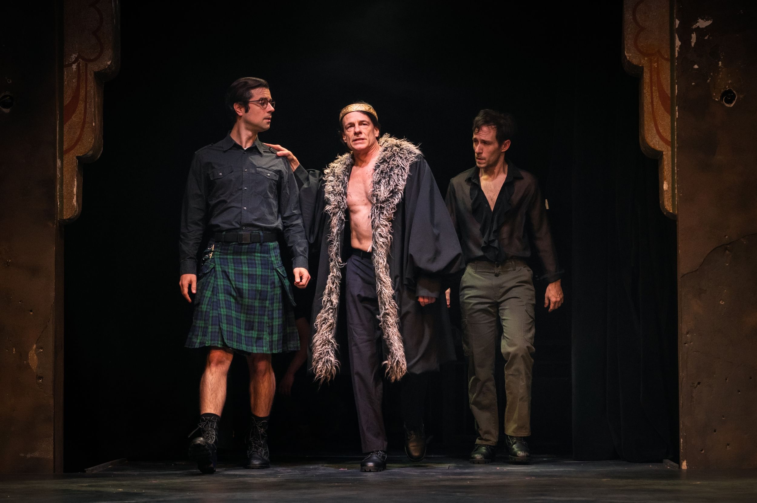 3 white male actors walk together, one in a kilt, one in a furred robe, and one in a drapey black open v-neck shirt.