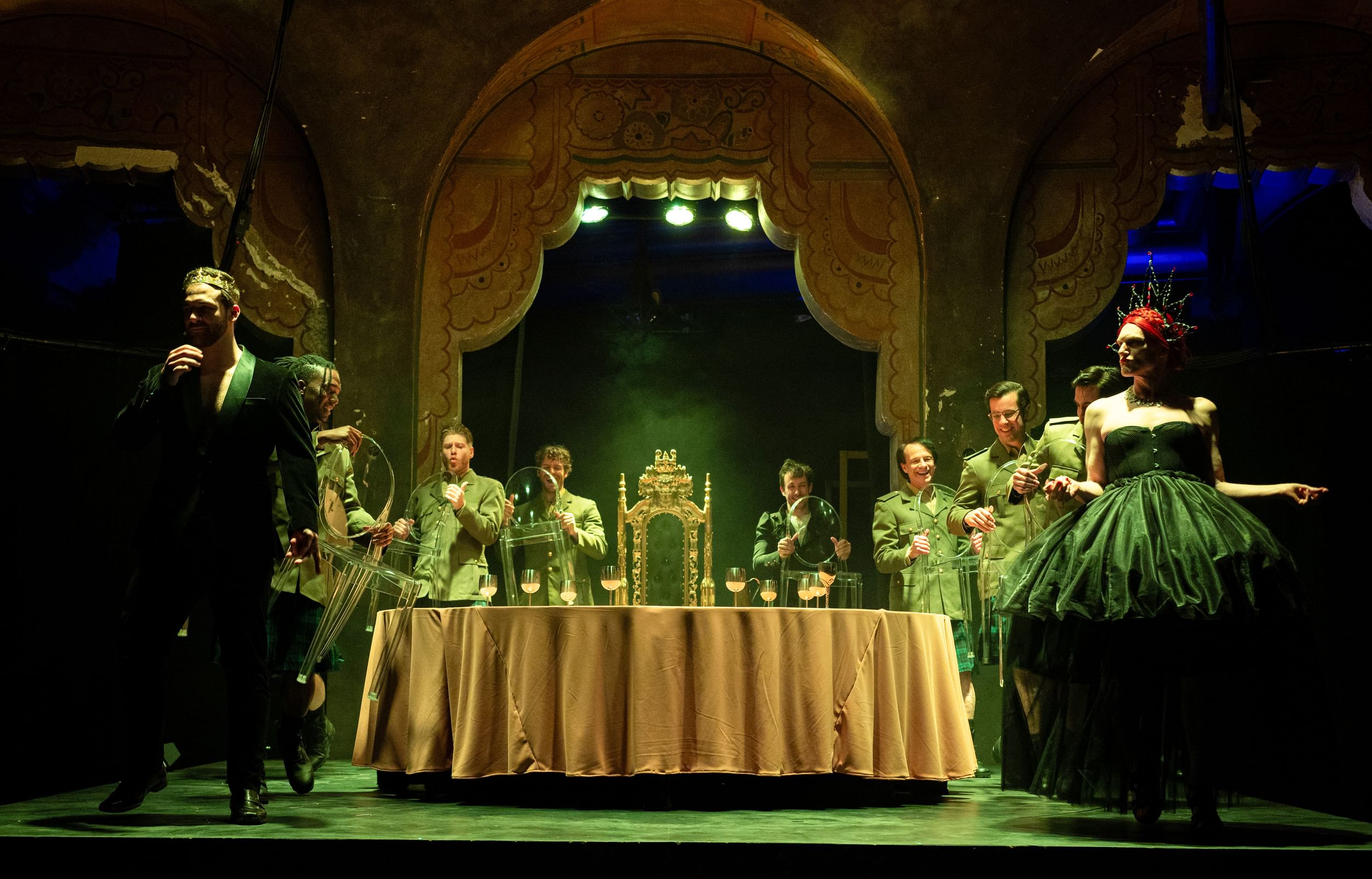 The cast, in vaguely military costumes, processes in a U-shape toward the camera, around a fancy cloth-covered dining table.